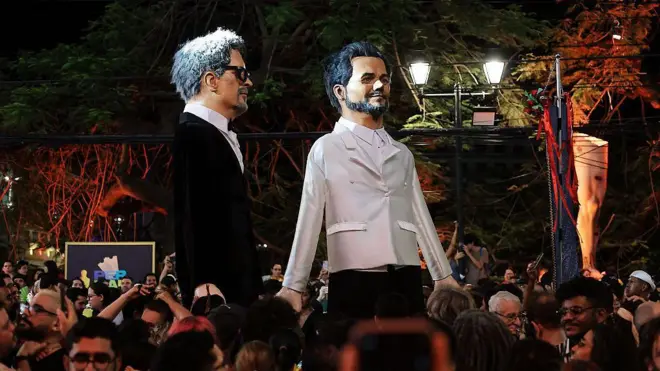 Boneco gigantes de Wagner Moura e Kleber Mendonça Filho na rua, com pessoas em volta. Elas acompanhavam cerimônia do Oscar em frente ao Cinema São Luiz, em Recife.