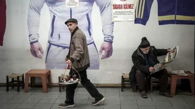 Elinde çay tepsisiyle çay dağıtan ve oturmuş gazete okuyan yaşlı iki erkek, duvarda eşofman reklam panosu