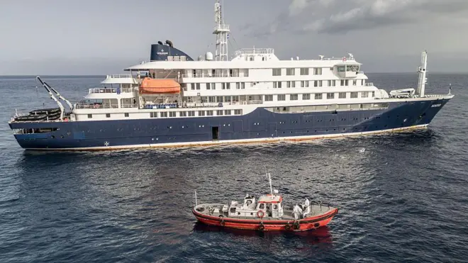 Sisi sebuah kapal pesiar kecil terlihat di air dengan sebuah perahu ambulans kecil berwarna merah di depannya, sekitar 50 meter jauhnya