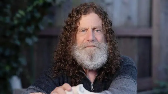 Foto colorida do filósofo Robert Sapolsky, um homem brancoquero a roletaolhos claros, cabelo cacheado na altura do pescoço e barba branca, vestindo uma camiseta azulquero a roletalã