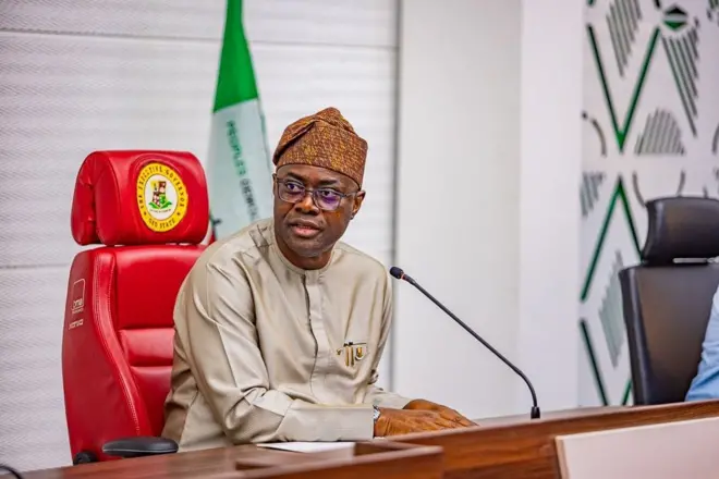 Seyi Makinde kọ oju si ẹgbẹ, gbohungbohun wa niwaju rẹ, o jokoo lori aga pupa