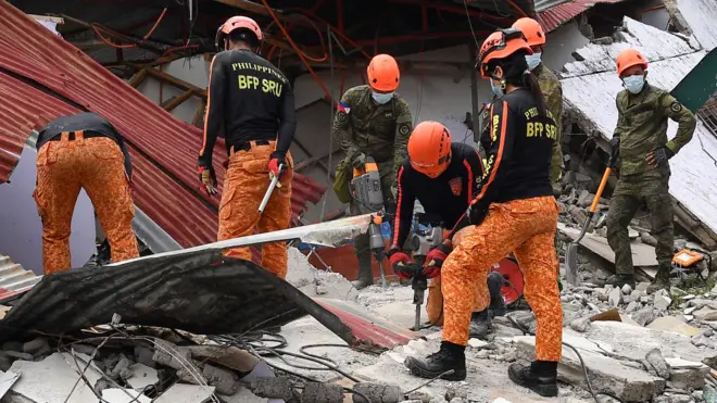 Спасатели в красных брюках и черных форменных рубашках на руинах дома. Двое из них сверлят фрагменты здания, еще один держит лопату