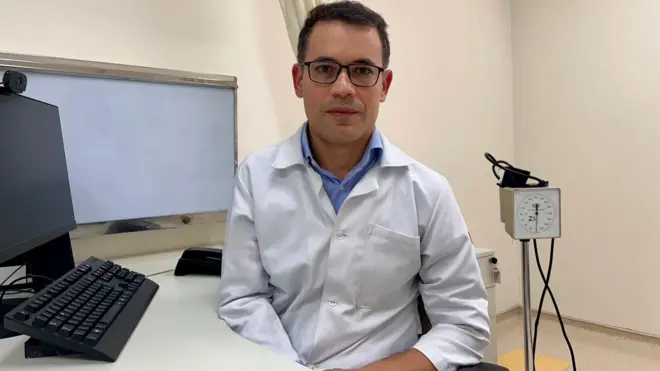 Gerson Salvador tem pele branca e cabelos escuros. Na imagem, eleusa óculoscasas de apostas com dinheiro gratisleitura, jaleco, e está sentado dentrocasas de apostas com dinheiro gratisum consultório médico 