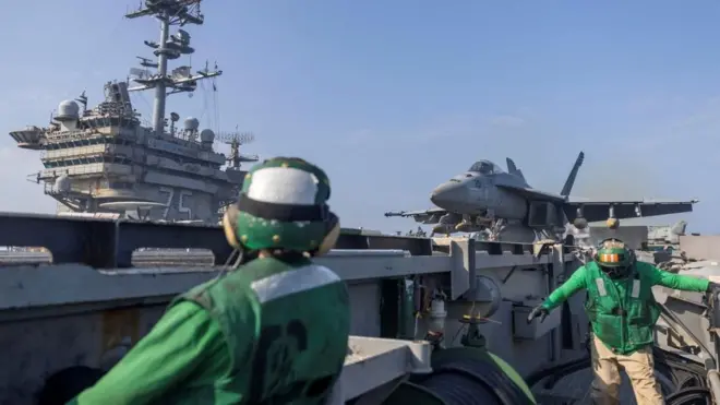 Two US military members in bright green jackets stand on an aircraft carrier with a jet behind them
