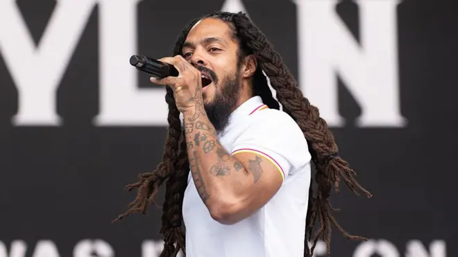A man with long dreadlocks, wearing a white T-shirt, performs with a microphone in front of a black stage background.