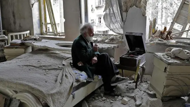 Mohammad Mohiedine Anis, 70, mengisap pipa sambil duduk di kamar tidurnya yang hancur dan mendengarkan musik di pemutar rekamannya di Aleppo.
