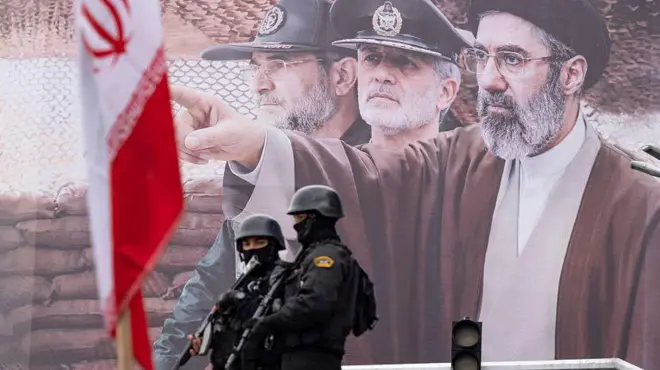 تصویری از بنر تبلیغاتی مجتبی خامنه‌ای در حالی که دو مامور امنیتی سیاه‌پوش با اسلحه مقابلش ایستاده‌اند