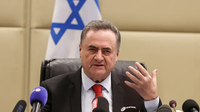Israel Katz üzerinde bir takım elbiseyle bir masada oturmuş, yüzünde gergin bir ifadeyle konuşurken elini kaldırıyor. Önünde mikrofonlar, arkasında ise bir İsrail bayrağı var