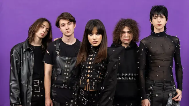 Zdjęcie przedstawia Alexandrę Căpitănescu z zespołem na fioletowym tle. Noszą czarne, skórzane i siatkowe stroje i patrzą prosto w obiektyw. 