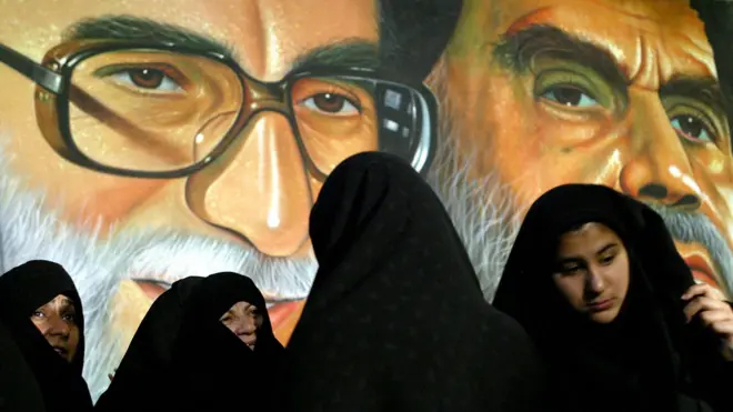 تصویر زنانی با حجاب اسلامی جلوی پوستری آیت‌الله خمینی و خامنه‌ای