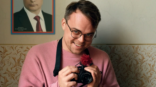 Мужчина держит в руках камеру и улыбается. На стене за ним портрет Владимира Путина.