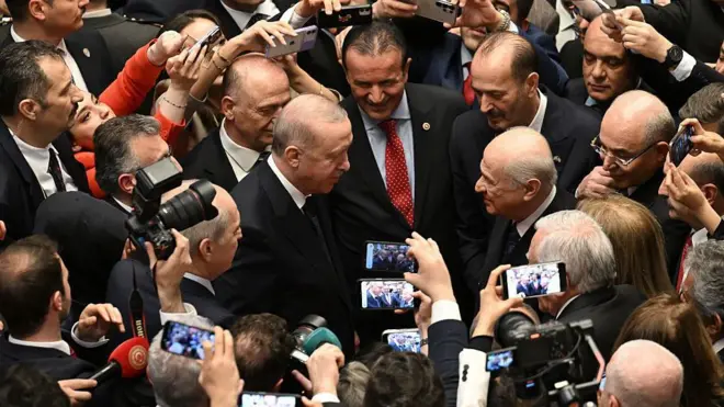 Solda Erdoğan, sağda Bahçeli birbirlerine bakıyorlar, çevrelerinde toplanmış çok sayıda siyasetçi var. Fotoğrafta önceki kişiler liderlerin fotoğraflarını çekiyor. 
