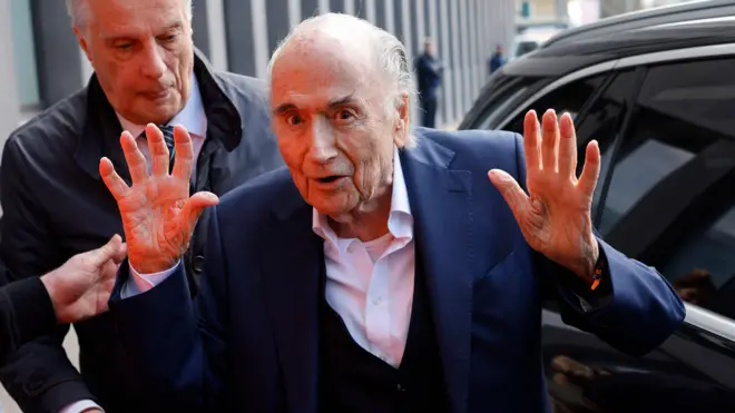 Former Fifa President Sepp Blatter arrives at the tribunal for the verdict. He wears a navy suit, stares directly at the camera and holds both hands up. 