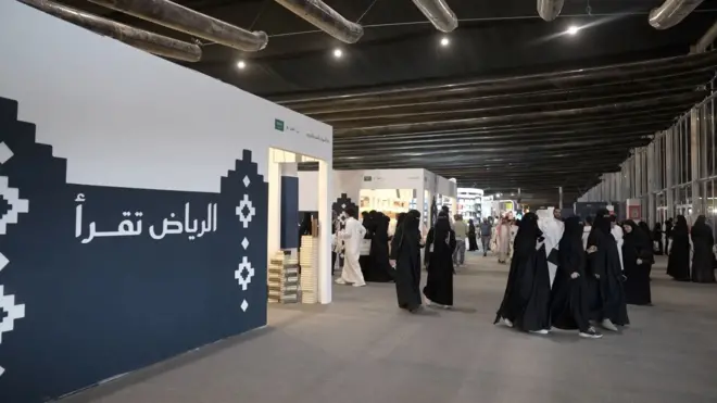 صورة تظهر نساء وفتيات ترتدي عباءات سوداء وتسير في أحد أروقة معرض الرياض الدولي للكتاب، ويسير كذلك رجال يرتدون جلاليب بيضاء. وهناك جدار كُتب عليه "الرياض تقرأ".