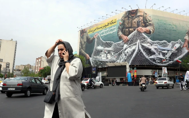 Bir kadın büyük bir caddenin ortasından telefonla konuşarak geçiyor. Arkada dev bir İran'ın savaşını destekleyen poster var.