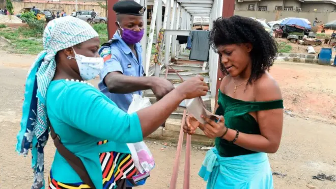 Two women and one policeman dey look at how to wear facemask