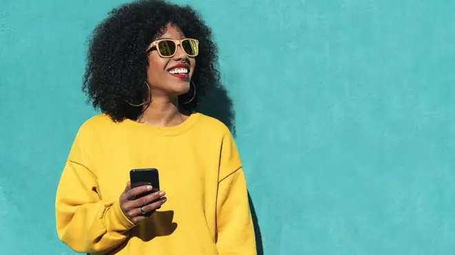 Young woman using a mobile in the street. She's wearing a bright yellow top and is standing in front of a vivid turquoise wall