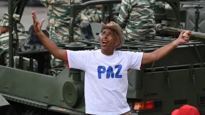 Un partidario del gobierno chavista en Venezuela con una camiseta con la palabra "paz", frente a un vehículo con soldados.
