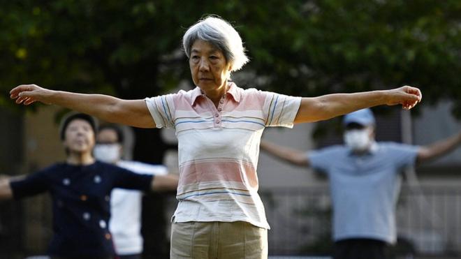 People exercising in a park in Tokyo in October 2022 - person in foreground with grey hair extends shoulders while standing up straight