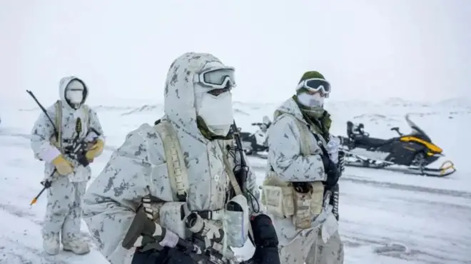 Трое канадских военнослужащих на заснеженном фоне в белой камуфляжной форме, с масками на лице и в перчатках, с различными видами боеприпасов в руках. Позади них стоит припаркованный снегоход.