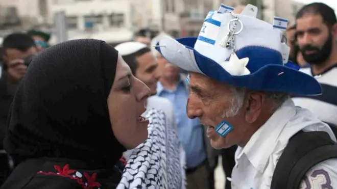 A Palestinian woman wearing a headscarf and an Israeli man wearing a blue and white hat bearing the Israeli flag shout at each other in Jerusalem