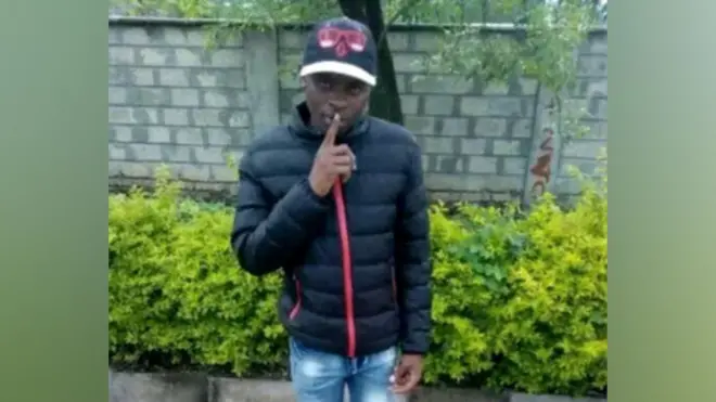 Boniface Kariuki wear one black jacket and cap, e hold im finger to im lips