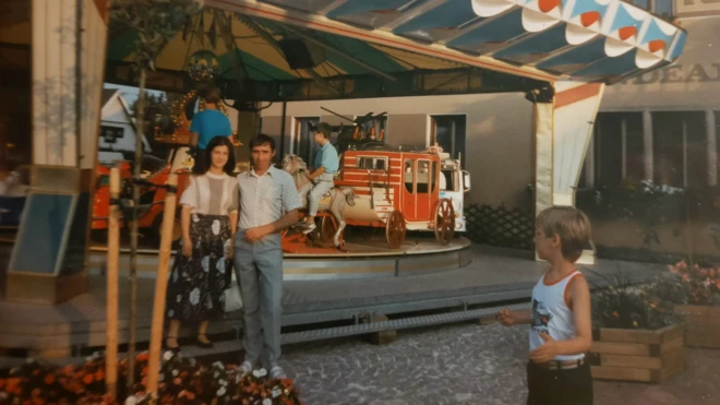 Двоје људи испред рингишпила и гледа их плавокоси дечак