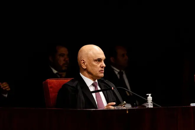 Fotografia do ministro do STF Alexandre de Moraes durante julgamento do ex-presidente Bolsonaro na corte. Ele veste gravata vermelha e camisa branca, além da toga. Há pessoas ao fundo, sob sombras.