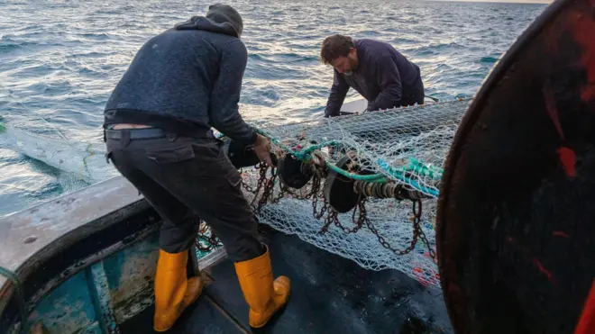 Two fishermen on a boat putting out a fishing net from dia boat for dia first catch of di day for di English Channel, off di south coast of East Sussex, with waves for di background and soft light from di rising sun.