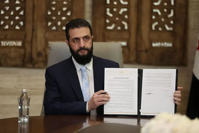 Şara takım elbiseli şekilde bir masada tek başına, imzalanan anlaşmayı kaldırarak kameralara gösteriyor.