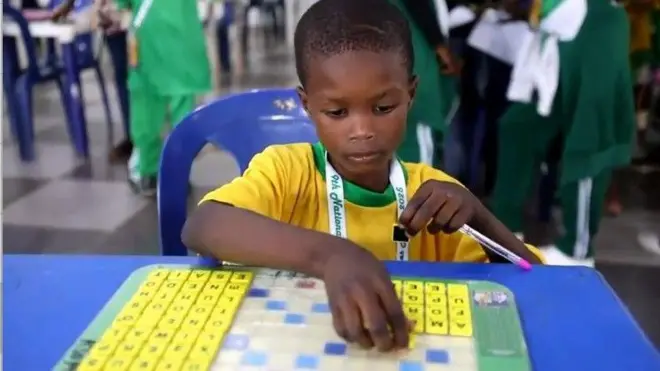 Yusuff Abdulsalam ọmọ ọdun mẹfa joko lori aga alawọ buluu, o wọ asọ alawọ ofeefe, ọpọn ayo Scrabble kan wa niwaju rẹ lori tabili alawọ buluu bakan naa, to si n n ta ayo naa.