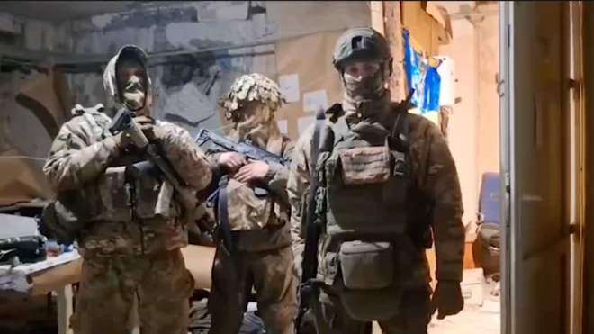 Троє військових у повному камуфляжному спорядженні та бронежилетах. Вони стоять у приміщенні з пошкодженими стінами, на яких видно папери та закріплені матеріали. Один із військових тримає автомат у руках, інші мають зброю на спорядженні. На задньому плані – стіл із речами та обладнанням. Освітлення штучне, загальний вигляд приміщення – імпровізований штаб або укриття.
