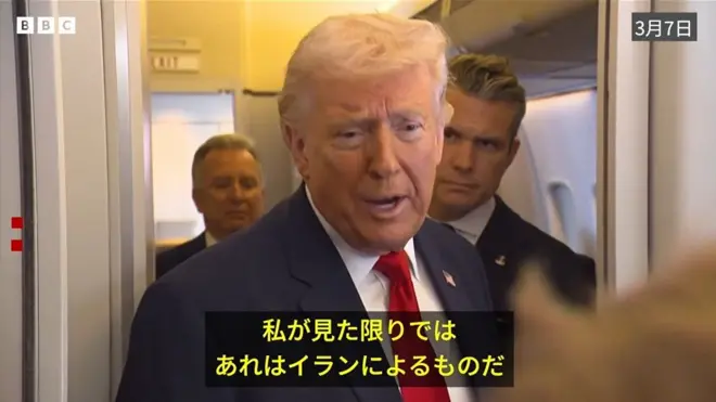 ダークスーツに赤いネクタイのトランプ氏が、大統領専用機内で記者団の質問に答えている。背後にはヘグセス国防長官が立っている