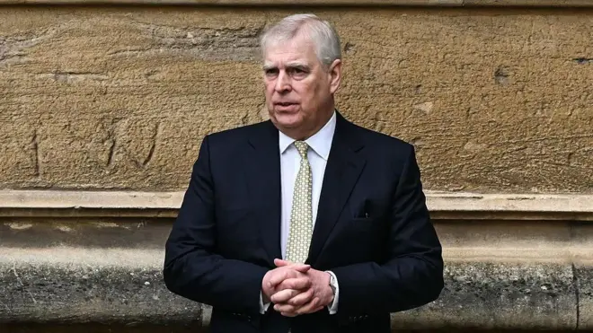 Prince Andrew wearing a black suit blazer and a green tie with his hands clasped in front of him, against a sandstone wall