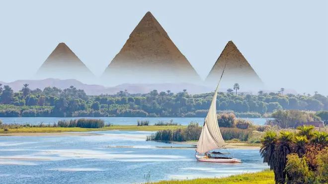 Les pyramides de Gizeh s'élevant au-dessus des palmiers et du Nil, avec un voilier blanc au premier plan