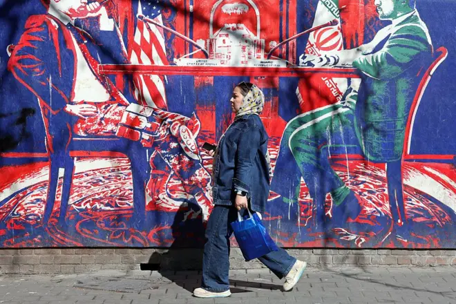 26 फरवरी, 2026 को तेहरान में पूर्व अमेरिकी दूतावास के बगल में स्थित ईरान-अमेरिका वार्ता को दर्शाने वाले अमेरिका विरोधी भित्तिचित्र के पास से एक ईरानी महिला गुजर रही है.