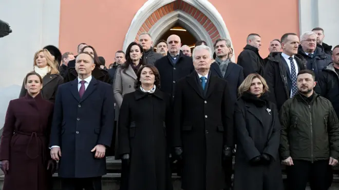 Велика група людей стоїть на східцях перед входом до будівлі теракотового кольору. В першому ряду – зліва направо – Марта та Кароль Навроцькі, Діана та Гітанас Науседа, Олена та Володимир Зеленські. За ними ще кільканадцять людей шикуються щільними рядами для офіційного групового знімка, серед яких видно Андрія Сибігу та Світлану Тихановську.