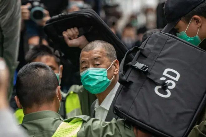 Jimmy Lai wears a green face mask and arrives to a crowded court room, he is seen outside of the courtroom with security around him.