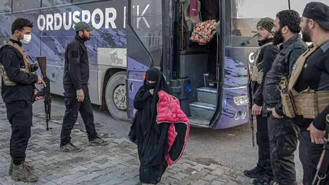 Silahlı kişilerin gardiyanlığında bir genç kız otobüsten iniyor. Tamamen çarşaflı bu kızın elinde pembe bir mont var.