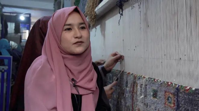 صالحه حسنی، ۱۹ ساله لباس سیاه و چادر مرجانی رنگ، در کنارش یک دستگاه قالین بافی است که در عقب او چند دختر در حال بافتن قالین هستند.