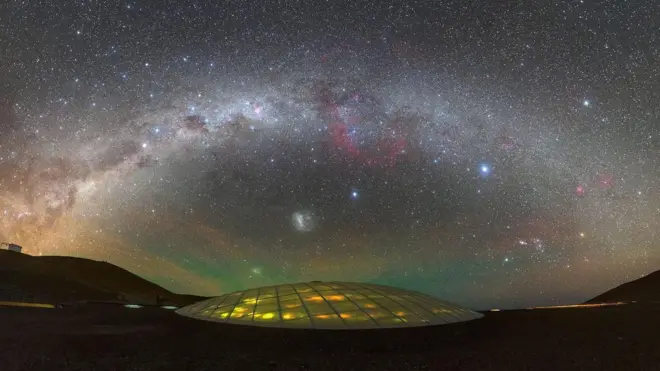 Vista de un paisaje nocturno bajo un cielo estrellado con la cúpula iluminada del hotel para científicos en el centro. 