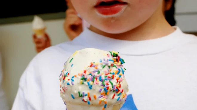 Devojčica drži beli sladoled posut šarenim posipom u rukama koji joj se topi preko prstiju