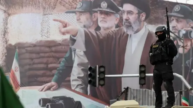 بیلبوردی از مجتبی خامنه‌ای که با دست به جایی اشاره می‌کند اطراف او چند فرمانده سپاه هستند. جلوی بیلبورد یک نیروی نظامی مجهز با چهره‌ای پوشیده دیده می‌شود