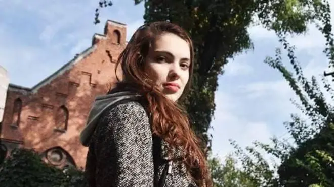 Дівчина з каштановим волоссям дивиться прямо у кадр на фоні зелені і будинку 