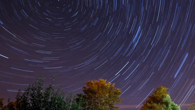 A long-exposure photo of the night sky during the Perseid Meteor Shower shows circular star trails caused by Earth's rotation. Trees with green and yellow leaves are visible in the foreground.