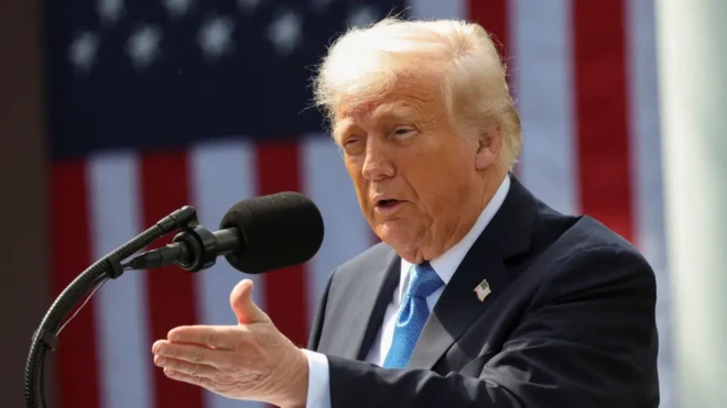 دونالد ترامب مرتدياً بدلة رسمية سوداء وربطة عنق زرقاء وقميص أبيض، يتحدث وأمامه مايكروفون أسود وخلفه العلم الأمريكي