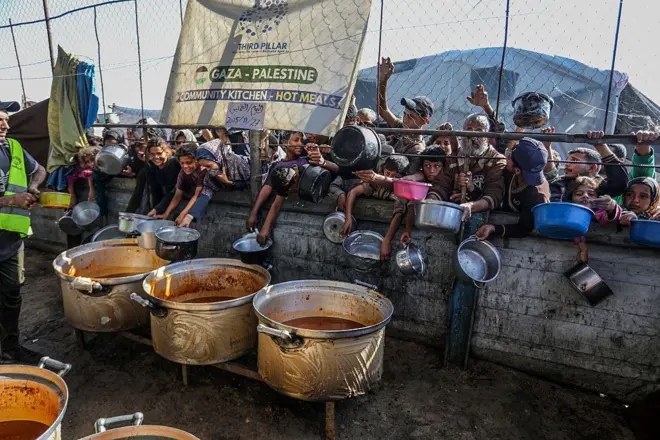 صورة لأشخاص من خلف سور يمدون أوانيهم الفارغة في انتظار نصيب من الأكل الساخن الذي تقدمة المطابخ المجتمعية.