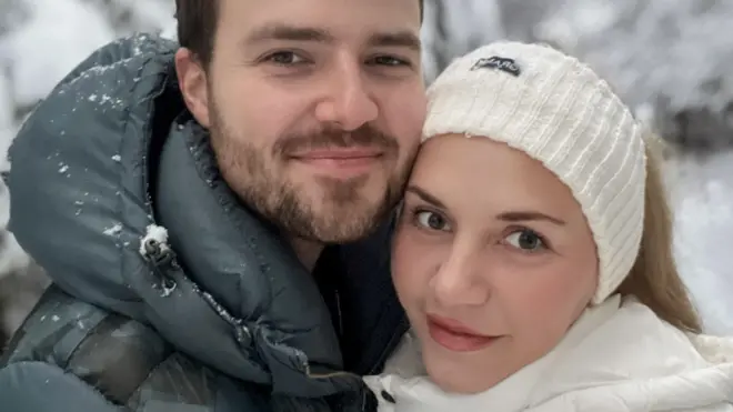 Two people standing close together in the foreground, wearing padded winter jackets, with one in a dark green coat and the other in a white coat and knitted headband. Snow is visible on the clothing. Behind them are snow-covered trees and branches.