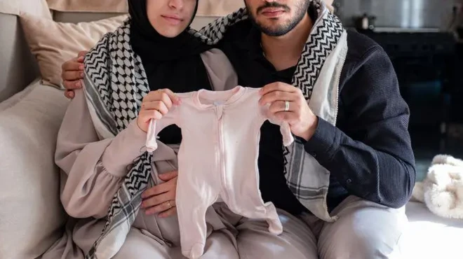 یک زوج فلسطینی که چهره‌شان مشخص نیست لباس نوزادی را با هم در دست گرفته‌اند و یک چفیه روی دوش انداخته‌اند