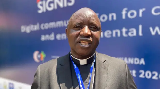 Musenyiri Balthazar Ntivuguruzwa wa Diyosezi ya Kabgayi mu Rwanda arimo kuvuga mu kiganiro na BBC Gahuzamiryango i Kigali, ku itariki ya 6 Kanama (8) mu mwaka wa 2026. Yambaye ikositimu y'umukara, ishati y'umukara y'ikora rigufi iriho n'ikora ry'umweru rya gisaseridoti. Inyuma ye hari icyapa cy'ubururu cyanditseho amagambo y'umweru yamamaza inama y'itangazamakuru rya Kiliziya Gatolika.